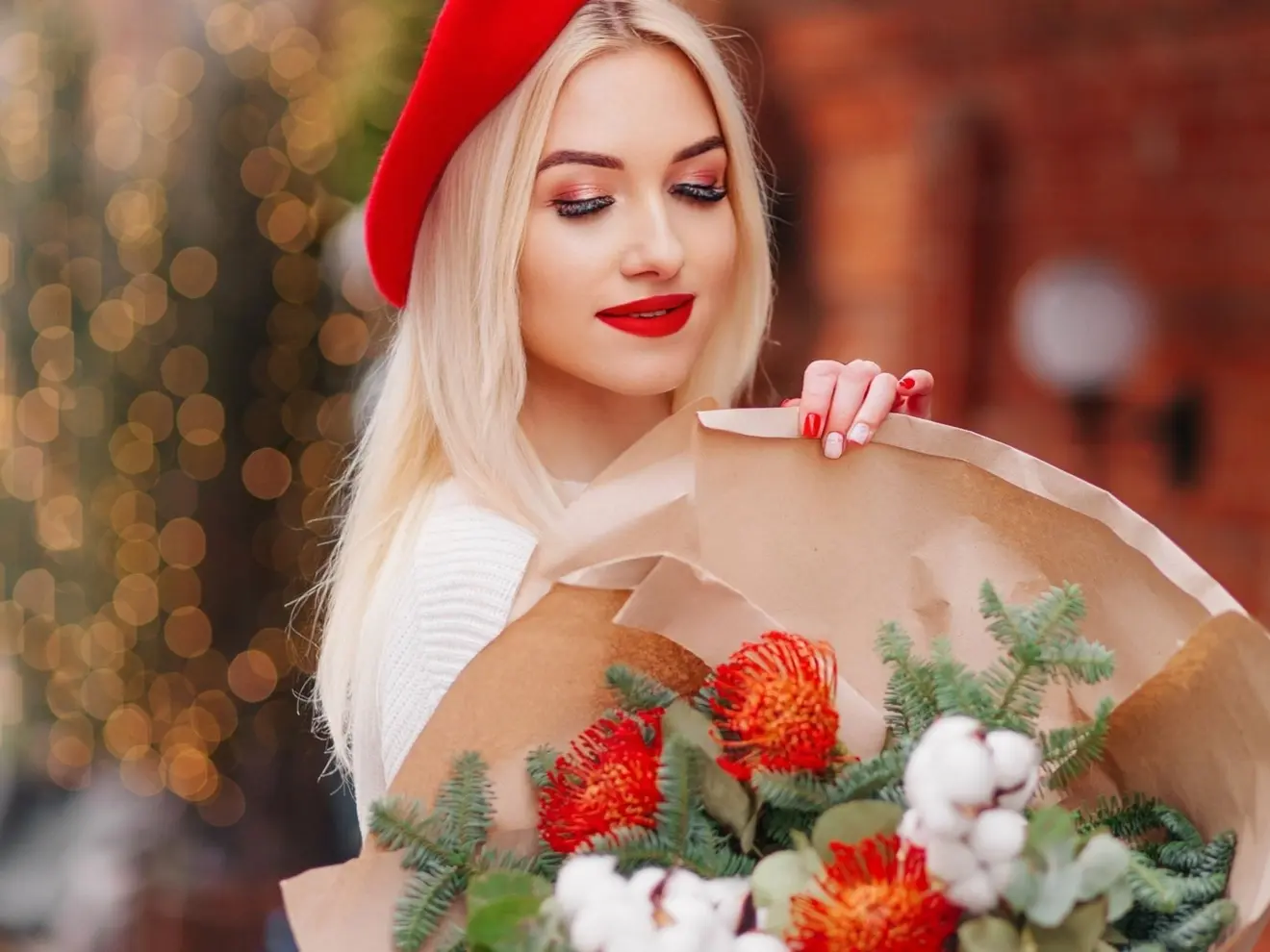 Woman in a red beret holding a holiday bouquet of red pincushion protea, pine, and cotton wrapped in kraft paper
